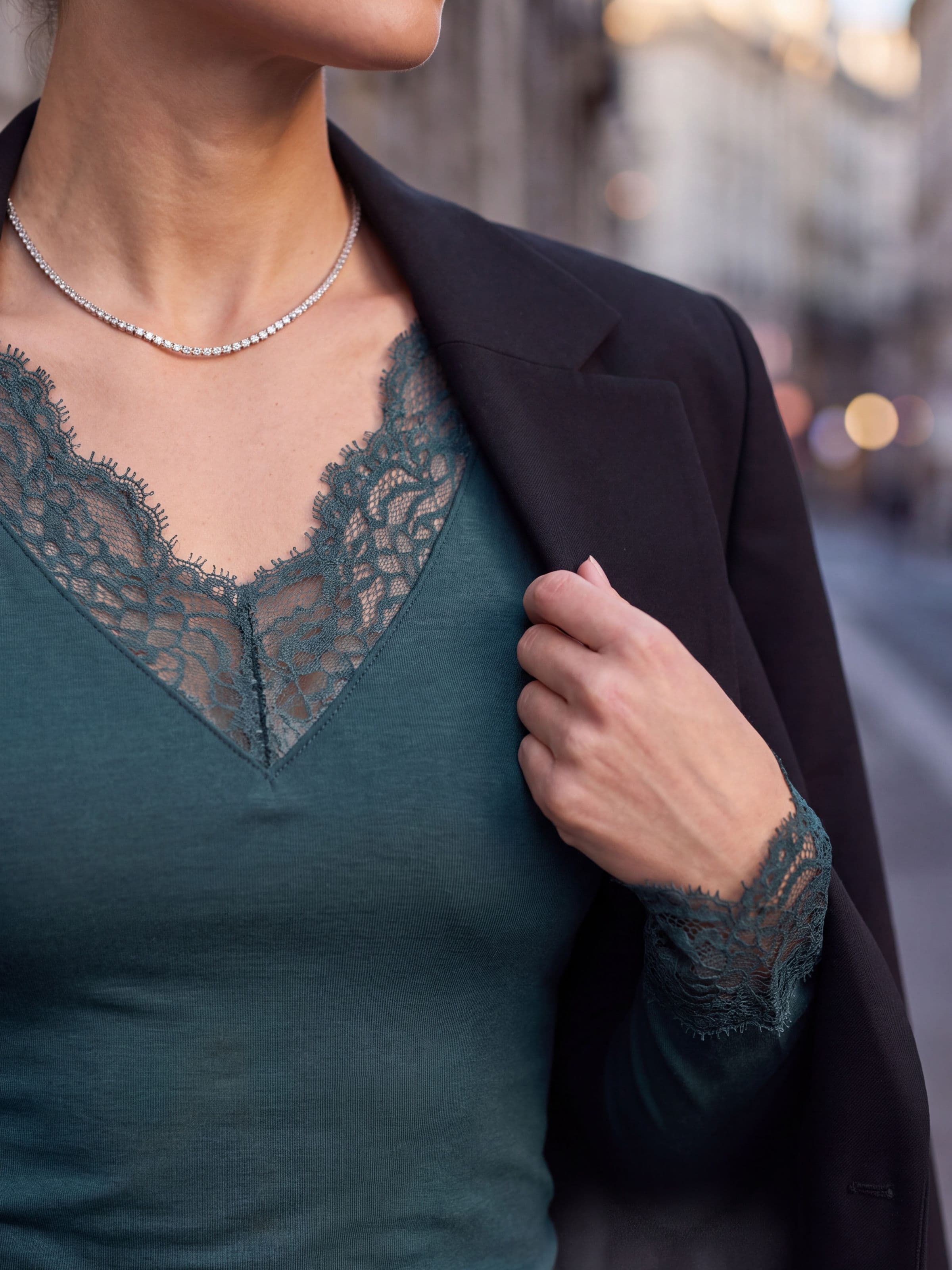 Detailaufnahme eines flaschengrünen Unterhemds mit V-Ausschnitt aus Spitze und Spitzenmanschette, getragen unter einem schwarzen Blazer mit einer Halskette
