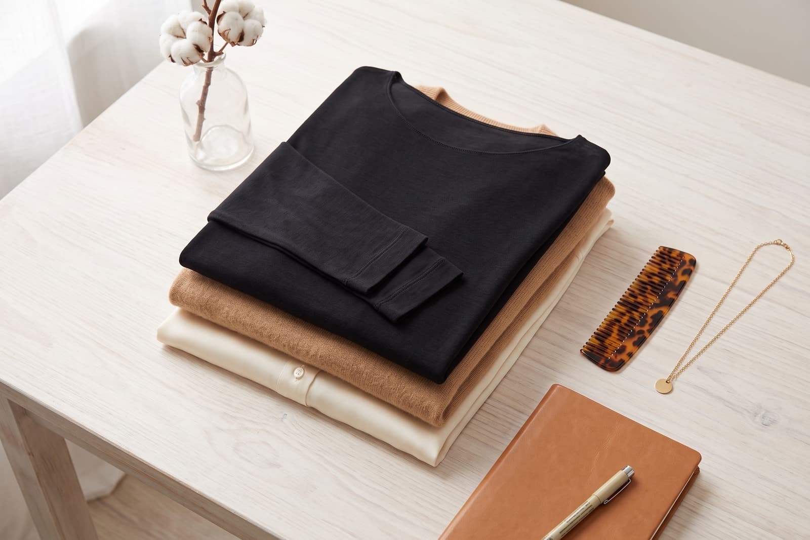 Schwarzes Damen-Unterhemd mit langen Ärmeln und Bootsausschnitt, gefaltet auf einem Kamelpullover und einer cremefarbenen Bluse