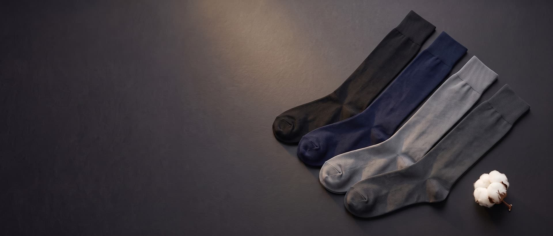 Vier Paar lange Socken aus Filo di Scozia vollstĂ€ndig ausgelegt, in Schwarz, Marineblau, Grau und Anthrazit, neben einer BaumwollblĂŒte