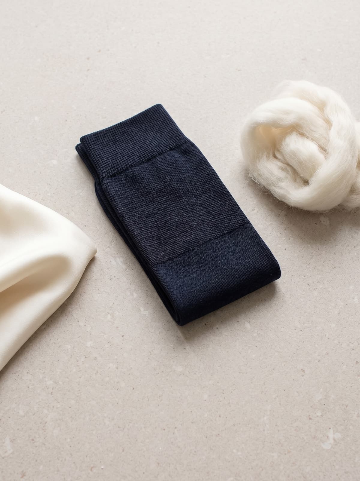 Navy blue over-the-calf sock in wool and silk next to a tuft of Merino wool and an ivory silk fabric