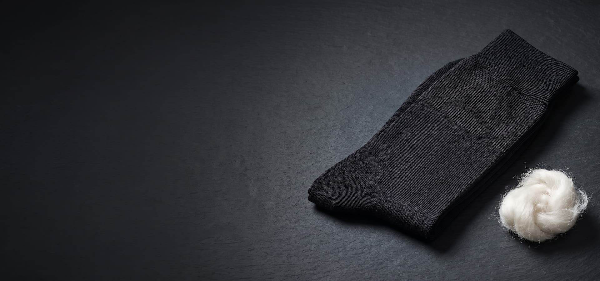 Black long wool and silk sock folded on dark slate next to a tuft of Merino wool