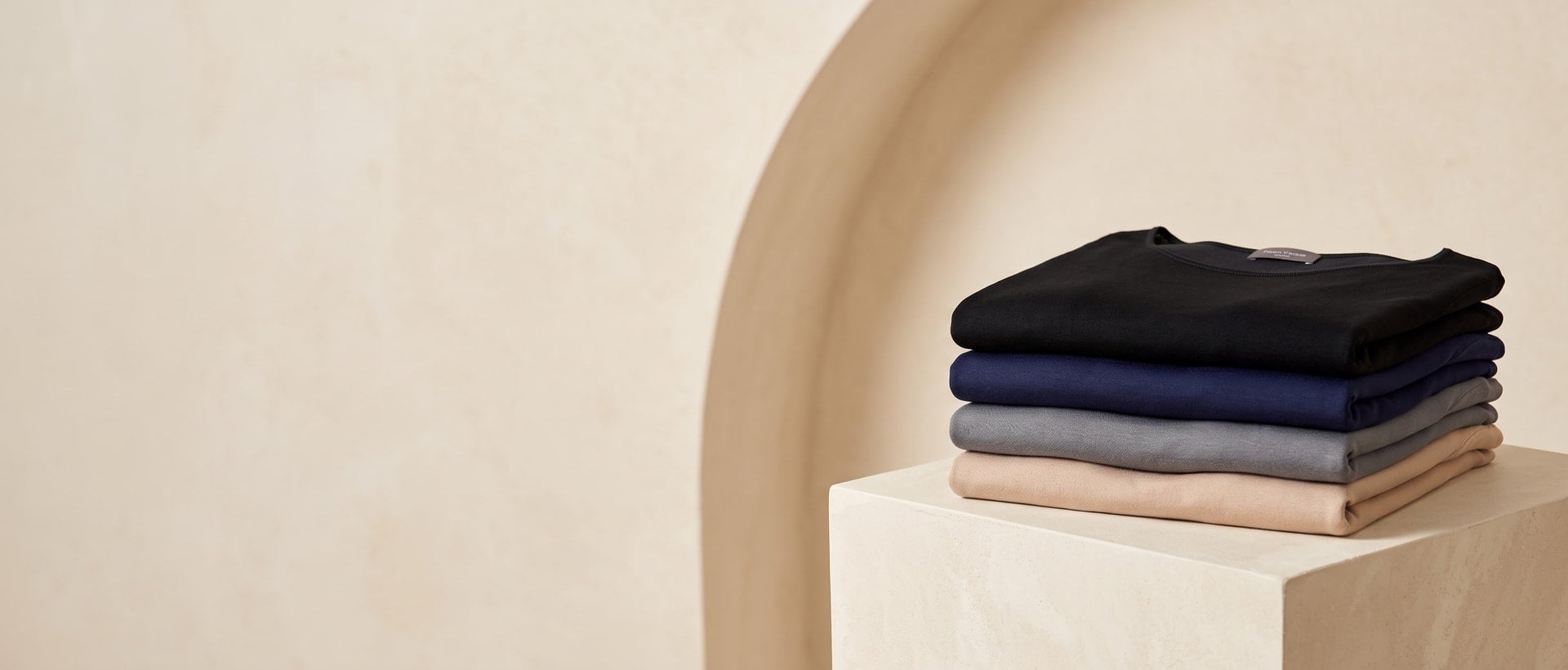 Camisetas dobladas en negro, azul marino, gris y beige sobre un plinto geométrico color crema
